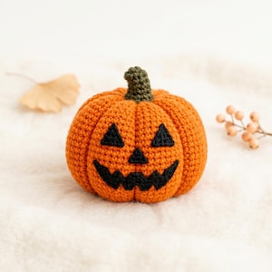 May include: A round, orange crocheted pumpkin with a dark green stem. The pumpkin features a black stitched jack-o'-lantern face with two triangular eyes, a small triangular nose, and a wide, jagged, toothy grin made of black yarn.