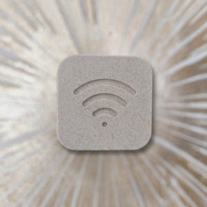 Può includere: Una placca quadrata color beige chiaro con angoli arrotondati che presenta un simbolo Wi-Fi in rilievo al centro. La superficie ha un aspetto punteggiato e strutturato, e l'icona è composta da tre linee curve sopra un piccolo punto.