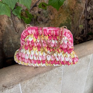Può includere: Una borsa all'uncinetto con un motivo multicolore di strisce di tessuto rosa, giallo e bianco intrecciate con un sottile filo metallico dorato. La borsa presenta un'apertura a telaio strutturata e una tracolla a catena in metallo dorato.