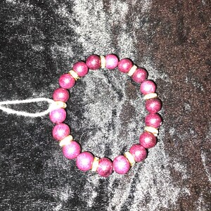 May include: A circular beaded bracelet featuring alternating dark magenta, textured spherical beads and smaller, metallic gold-toned spacer beads. The bracelet is strung on an elastic cord and includes a small white tag attached to one side.