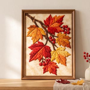 May include: A framed wall art piece featuring a three-dimensional fabric collage of autumn maple leaves in shades of red, orange, and yellow, arranged on a branch with small red berries against a textured cream-colored background.