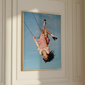 Könnte beinhalten: Ein gerahmter Kunstdruck, der eine Person in einem rosa Kleid, weißen Socken und schwarzen Schuhen zeigt, die kopfüber auf einer Metallkettenschaukel vor einem klaren blauen Himmel schwingt. Das Bild ist in einem dünnen Goldrahmen gefasst.