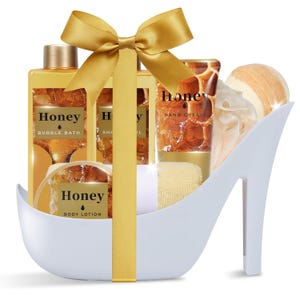 May include: A white high-heeled shoe-shaped container holding honey-themed bath products including bubble bath, shower gel, hand cream, body lotion, a bath sponge, and a bath bomb, all tied together with a large gold ribbon and bow.
