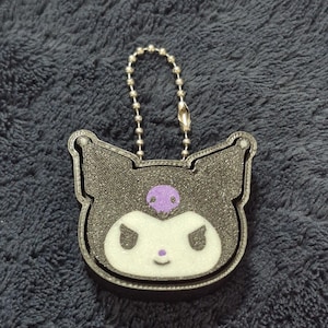 May include: A black and white character keychain featuring a jester-style hat with a purple skull emblem. The character has a white face with pointed ears, a small purple nose, and a neutral expression. A silver metal ball chain is attached to the top.