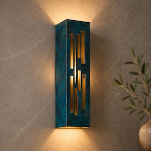 May include: A rectangular wall sconce with a weathered teal and blue patina finish. The fixture features vertical, narrow rectangular cutouts that allow warm light to project outward, creating a glow above and below the unit on the wall.