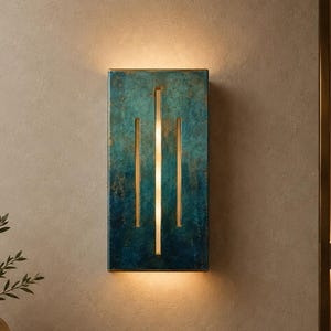 May include: A rectangular wall sconce featuring a textured, mottled finish in shades of deep blue and teal with gold accents. The fixture has three vertical cutouts that emit a warm, soft glow, illuminating the wall above and below the unit.