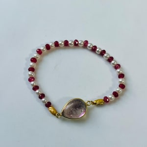 Op de afbeelding: Een kralenarmband met afwisselend witte parels en gefacetteerde donkerrode kralen. De armband heeft een centrale druppelvormige lichtroze edelsteen in een goudkleurige metalen zetting, geflankeerd door twee langwerpige goudkleurige kralen.