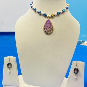 Op de afbeelding: Een ketting- en oorbellenset bestaande uit een choker met witte kralen en iriserende druppelvormige kralen, met in het midden een grote, goudomrande, paars en goudkleurige getextureerde druppelhanger. De bijpassende oorbellen hebben ovale stenen.