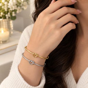 May include: Two thin metal bangle bracelets, one in a polished gold tone and one in a polished silver tone, each featuring a central knot design. The bracelets are worn together on the wrist, showcasing a smooth, reflective finish and a minimalist circular shape.