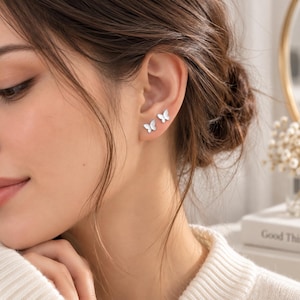 May include: A pair of small, silver-toned butterfly stud earrings worn on an earlobe. The earrings feature a simple, smooth silhouette in the shape of a butterfly with wings spread, providing a subtle and clean look for everyday wear.