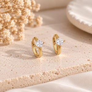 May include: A pair of gold hoop earrings, each featuring a single square-cut clear gemstone set in the center. The earrings have a polished metallic finish and a hinged closure mechanism, showcasing a simple and refined design for everyday wear.