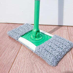 May include: A gray crocheted mop cover fits over a green and white mop head. The mop head is attached to a green and silver mop handle.