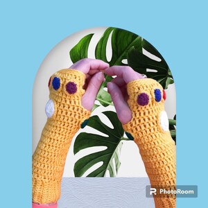 Pode incluir: Um par de luvas sem dedos de crochê amarelo com pontos coloridos no dorso da mão, que se assemelham à Manopla do Infinito do Universo Cinematográfico Marvel.