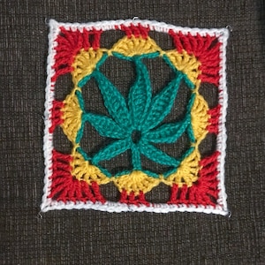 以下が含まれることがあります： 中央に緑色のマリファナの葉のデザインが施された、かぎ針編みの正方形。葉は黄色と赤色のかぎ針編みの円に囲まれています。