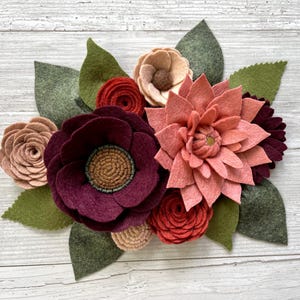 May include: A handcrafted felt flower arrangement featuring various blooms in shades of burgundy, peach, and red, with green felt leaves. The arrangement includes a large dahlia, several roses, and other flower types, all set against a light wood background.