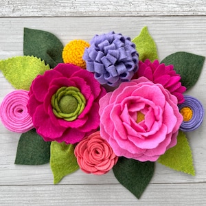 May include: A felt flower bouquet with pink, purple, and green felt flowers. The bouquet is arranged on a white wooden background.