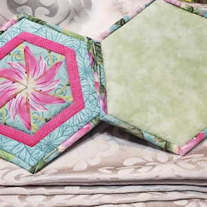 May include: Two hexagonal quilted fabric pads. One pad features a central pink flower motif on a light blue background with a bright pink border. The other pad has a solid, mottled light green fabric surface. Both items have matching edges.