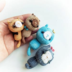 May include: Four small crocheted cat figurines in orange, tan, blue, and gray. Each cat features a round head, a white muzzle, black bead eyes, and thin whiskers. The tan cat has a blue bow, and the gray cat has a small pink bow on its head.