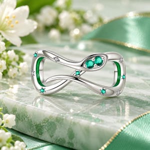 May include: A silver ring shaped like an infinity symbol with a snake head design. The ring features two round green stones for the eyes, accented with smaller round green and clear stones embedded along the polished silver band surface.