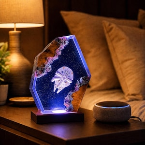 May include: A hexagonal resin lamp featuring a miniature Millennium Falcon spaceship embedded within a deep blue, starry background. The piece is framed by natural wood edges and sits on a dark rectangular wooden base with an integrated light.