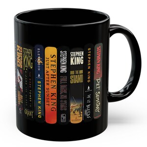May include: A black ceramic mug featuring a wrap-around graphic of various Stephen King book spines. The visible titles include Billy Summers, The Dark Tower IV, Elevation, Just After Sunset, Full Dark No Stars, The Stand, If It Bleeds, and Pet Sematary.