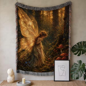 May include: A woven throw blanket featuring a detailed illustration of a fairy with large golden wings standing in a dark forest among mushrooms. The blanket has a fringed edge with multicolored threads and a warm, golden-toned color palette.