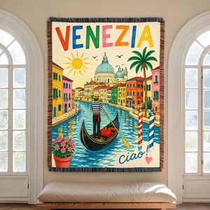 May include: A woven throw blanket featuring a colorful illustration of Venice with the text VENEZIA at the top and Ciao! at the bottom. The scene depicts a gondolier in a striped shirt rowing a gondola through a canal lined with buildings.
