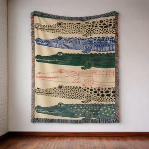 May include: A woven throw blanket featuring a cream-colored background with six horizontal rows of stylized alligator illustrations in varying colors including black, blue, green, and red, finished with a decorative fringed edge on all sides.