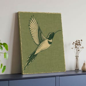 May include: A framed wall art print featuring a stylized illustration of a hummingbird in flight. The bird is rendered in cream and dark green tones against a textured olive green background, enclosed by a thin, decorative gold-colored border.