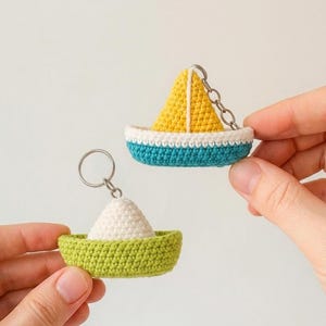 May include: Two crochet boat keychains. One features a yellow sail on a teal and white hull. The other has a white sail on a light green hull. Both include a silver metal keyring. The text Sugar Riot Stitches is visible in the bottom corner.
