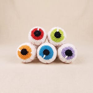 Puede incluir: Un conjunto de cinco globos oculares esféricos de ganchillo dispuestos en forma de pirámide. Cada ojo presenta una esclerótica blanca con venas rojas bordadas, una pupila negra circular y un iris de color rojo, verde, naranja, azul o morado.