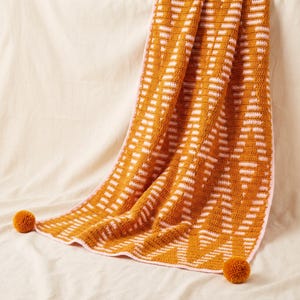 Könnte beinhalten: Eine rechteckige Häkeldecke mit einem geometrischen Muster in Senfgelb und Hellrosa. Die Decke ist mit einer hellrosa Umrandung versehen und hat zwei senfgelbe Bommeln, die an den unteren Ecken des Textilstücks befestigt sind.