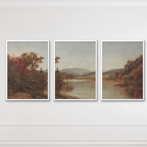 May include: A set of three framed landscape paintings featuring autumn scenery with trees in shades of red, orange, and brown, a calm river reflecting the sky, and distant mountains under a soft, overcast sky, all enclosed in white frames.