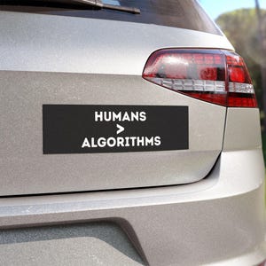 以下が含まれることがあります： HUMANS > ALGORITHMSという白い文字が書かれた長方形の黒いバンパーステッカー。ステッカーは車両のシルバーのトランクに貼られており、リアウィンドウの下、赤いテールライトアセンブリの左側に水平に配置されています。
