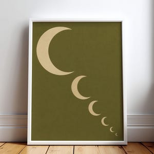 May include: A framed wall art print featuring a solid olive green background with a series of six cream-colored crescent moons arranged in a diagonal line, decreasing in size from the top left corner toward the bottom right corner of the frame.