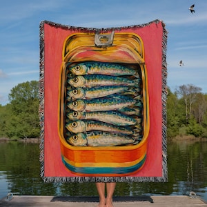 May include: A woven throw blanket featuring a vibrant illustration of an open tin can filled with stacked sardines. The background is a solid pink, and the can is yellow and orange. The blanket has a decorative fringed edge on all four sides.