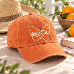 May include: An orange baseball cap featuring an embroidered butterfly graphic on the front panel. The butterfly design is outlined in white and contains the word Dolly in white cursive script across the center of the wings.