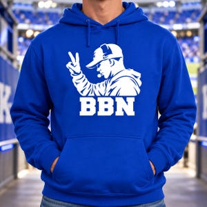 May include: A royal blue hooded sweatshirt featuring a white graphic print of a person wearing a baseball cap and making a peace sign gesture. Below the graphic, the text BBN is printed in a bold, white, block-style font.