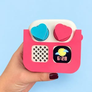 May include: A square-shaped digital clock with a bright pink frame and a white top section. It features two heart-shaped buttons in blue and pink, a circular speaker grille with small holes, and a digital display showing a yellow sun icon and 6:28.