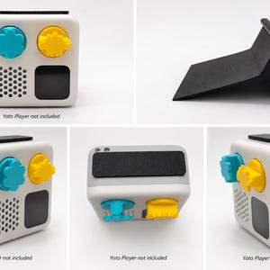 Puede incluir: Un soporte inclinado negro para un Yoto Player, mostrado desde múltiples perspectivas. El soporte cuenta con una base plana y un respaldo, diseñado para sostener el reproductor de audio blanco de forma cuadrada con sus perillas azules y amarillas.