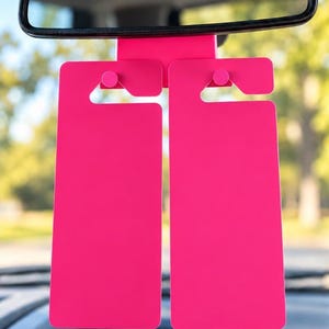 May include: A pair of bright hot pink rectangular car mirror tags hanging from a central mount. Each tag features a smooth, solid-coloured surface with a notched top section designed to hook securely onto a vehicle rearview mirror stem.