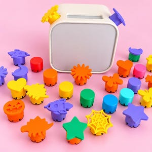 Op de afbeelding: Een wit vierkant apparaat met een leeg scherm, omringd door een verzameling kleurrijke plastic stempels in diverse vormen, waaronder sterren, harten, pompoenen, spinnen, vlinders en geometrische figuren in rood, oranje, geel, groen, blauw en paars.