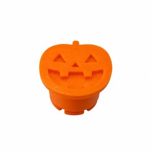 May include: A bright orange jack-o'-lantern topper featuring a carved face with two triangular eyes, a small triangular nose, and a wide, toothy grin. The object has a flat, circular top surface and a cylindrical base with small notches.