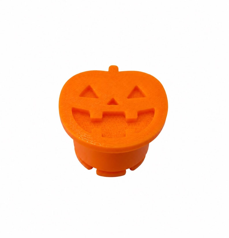 May include: A bright orange jack-o'-lantern topper featuring a carved face with two triangular eyes, a small triangular nose, and a wide, toothy grin. The object has a flat, circular top surface and a cylindrical base with small notches.