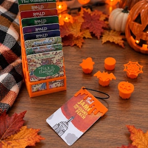 May include: An orange plastic tiered display stand holding several rectangular book-themed cards, including titles by Roald Dahl and Enid Blyton. Nearby are five small orange plastic Halloween-themed toppers and a James and the Giant Peach card.