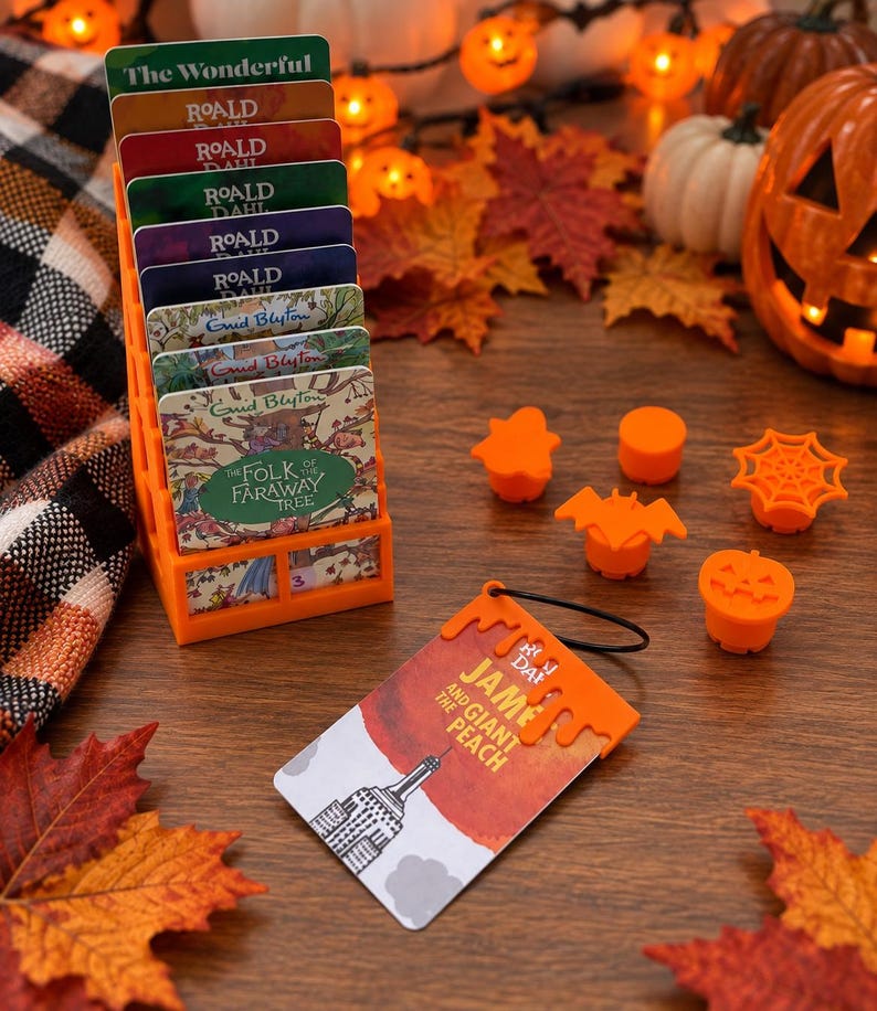May include: An orange plastic tiered display stand holding several rectangular book-themed cards, including titles by Roald Dahl and Enid Blyton. Nearby are five small orange plastic Halloween-themed toppers and a James and the Giant Peach card.