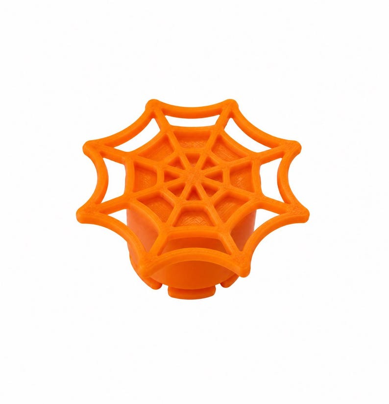 May include: A bright orange knob featuring a raised spider web design on its circular top surface. The object has a solid cylindrical base with a textured exterior and a flat, webbed pattern extending outward from the center to the edges.