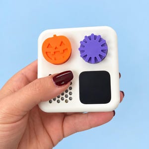 Puede incluir: Un juguete sensorial cuadrado de color blanco con cuatro elementos táctiles distintos: un botón naranja con forma de calabaza, un engranaje giratorio morado, una rejilla de pequeños huecos circulares y una superficie rectangular negra lisa.