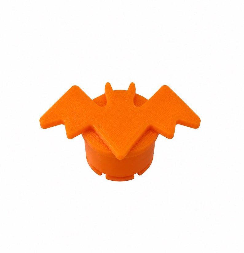 May include: A bright orange bottle stopper featuring a flat, stylized bat silhouette attached to the top. The cylindrical base has a textured surface with visible horizontal lines, and the bat has pointed ears and outstretched wings.