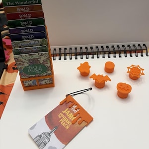 May include: An orange plastic card holder containing several book-themed cards, five small orange plastic toppers in shapes including a bat, pumpkin, and spiderweb, and one orange plastic book tag attached to a card titled James and the Giant Peach.